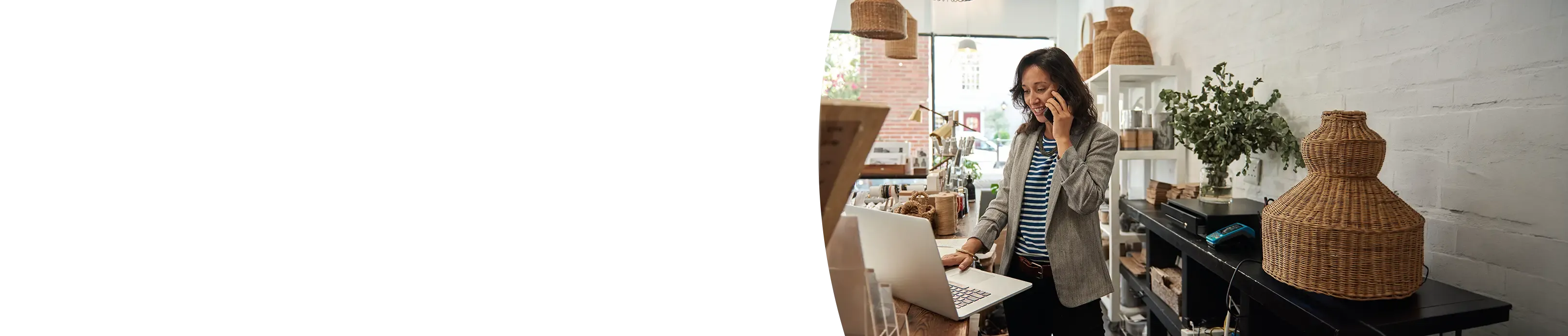 A woman business owner talks on a smartphone while using a laptop behind the counter of a boutique shop filled with home decor.